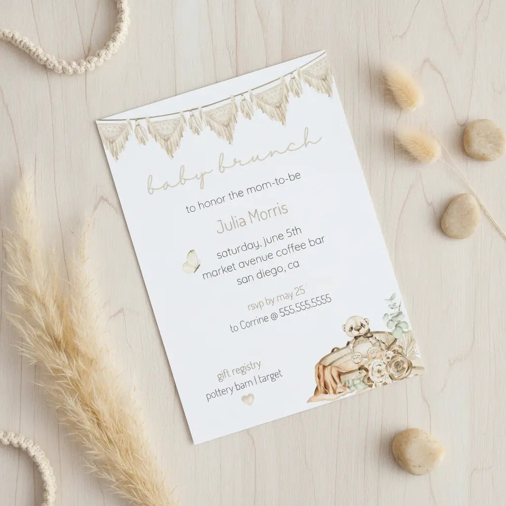 Customized boho baby shower invitation card with gender neutral teddy bear, macramé, and greenery on a wooden surface with decorative elements.
