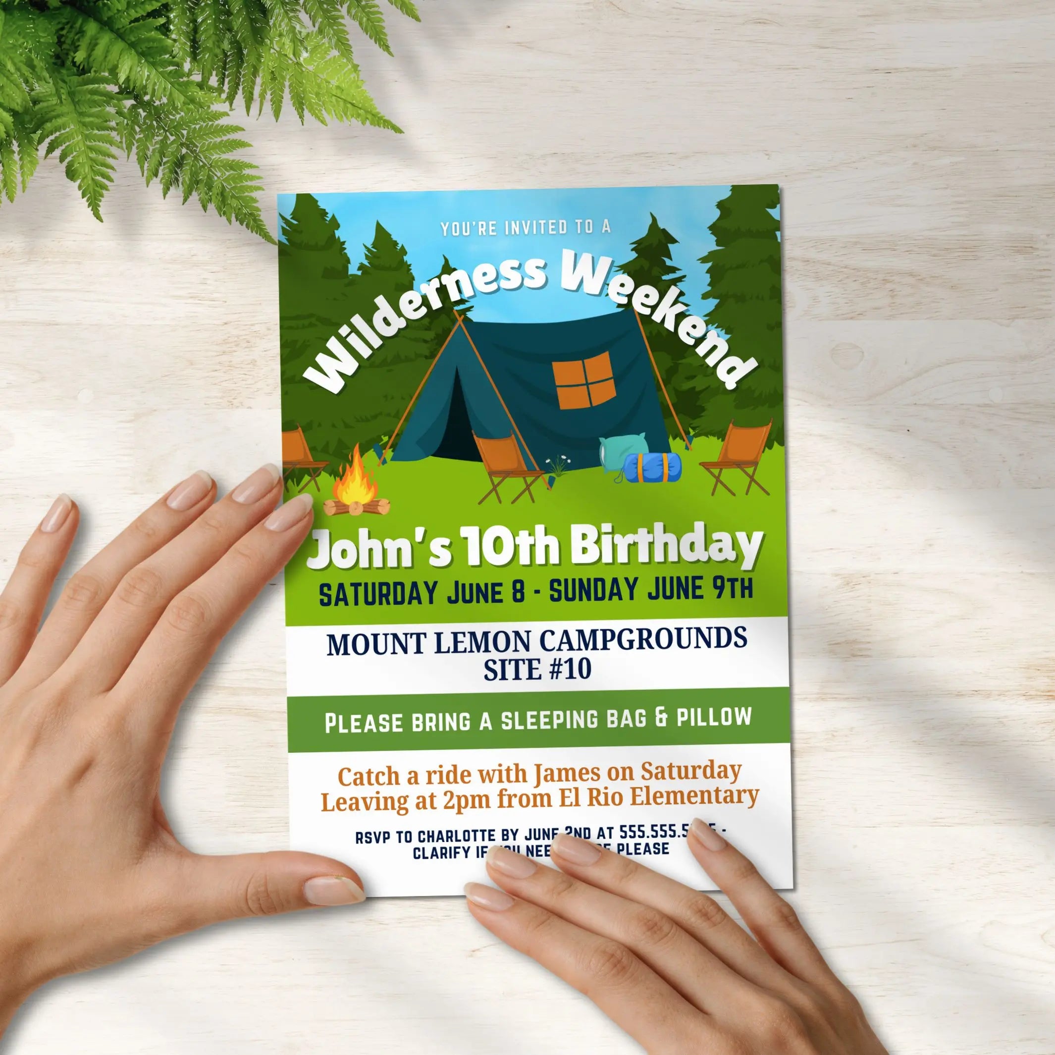 Invitation card for a wilderness weekend birthday party held by hands on a wooden surface with ferns.