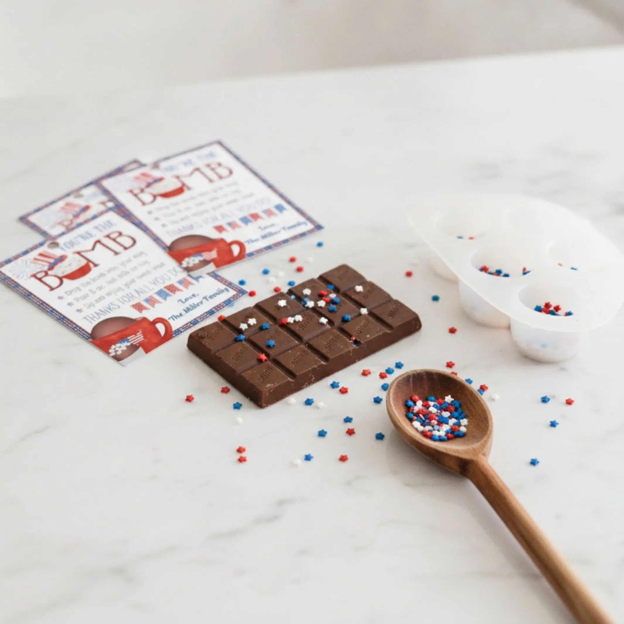 Chocolate bar with sprinkles, wooden spoon, and customized 'You're the Bomb' gift tag card on a white surface