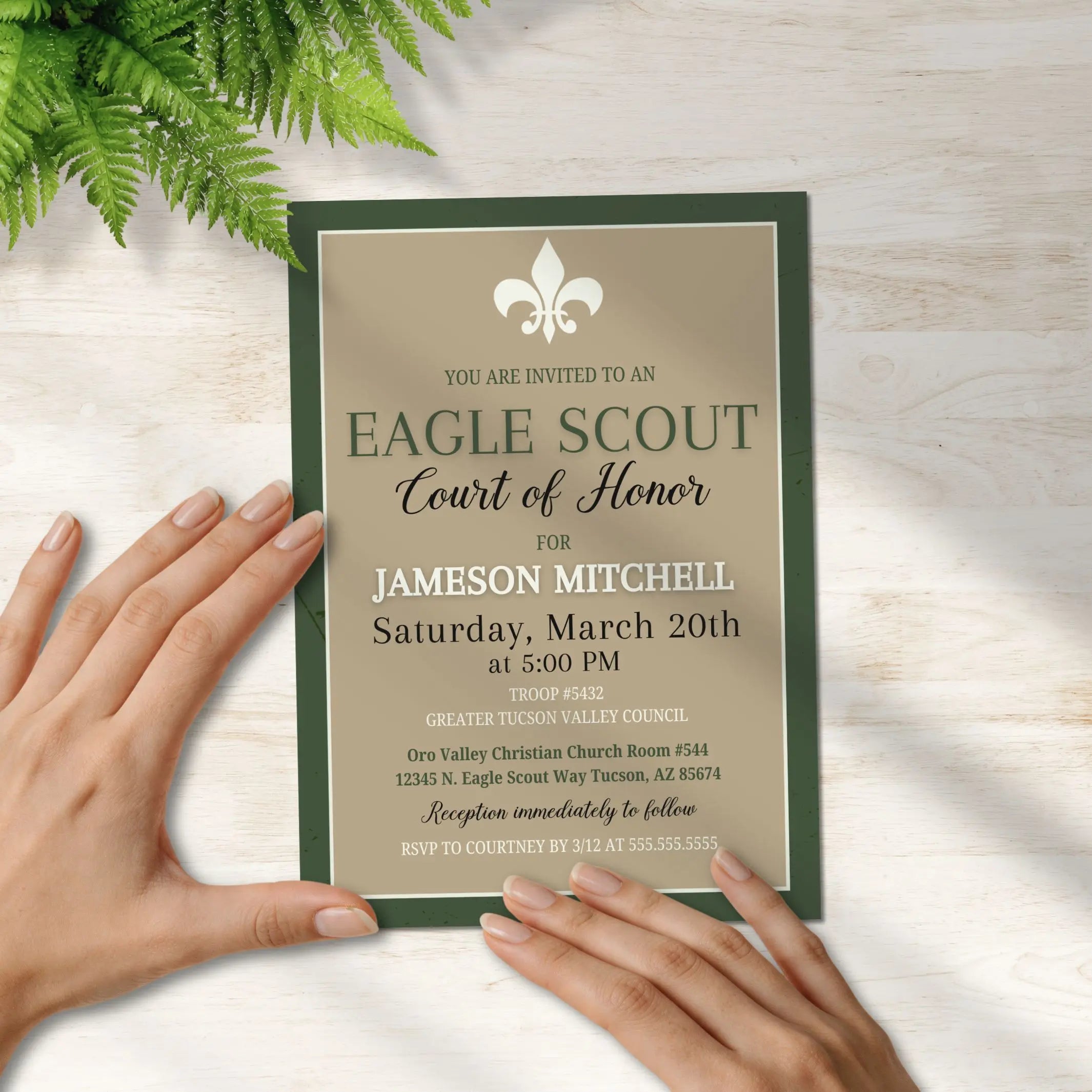 Eagle Scout Court of Honor invitation held by hands on a light wooden surface with greenery.