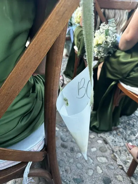 A close-up outdoor shot of a white vellum cone aisle marker with green elegant stems, held in place by a small green ribbon. The cone is decorated with a small green leaf and has the letters 'BC' written on it.