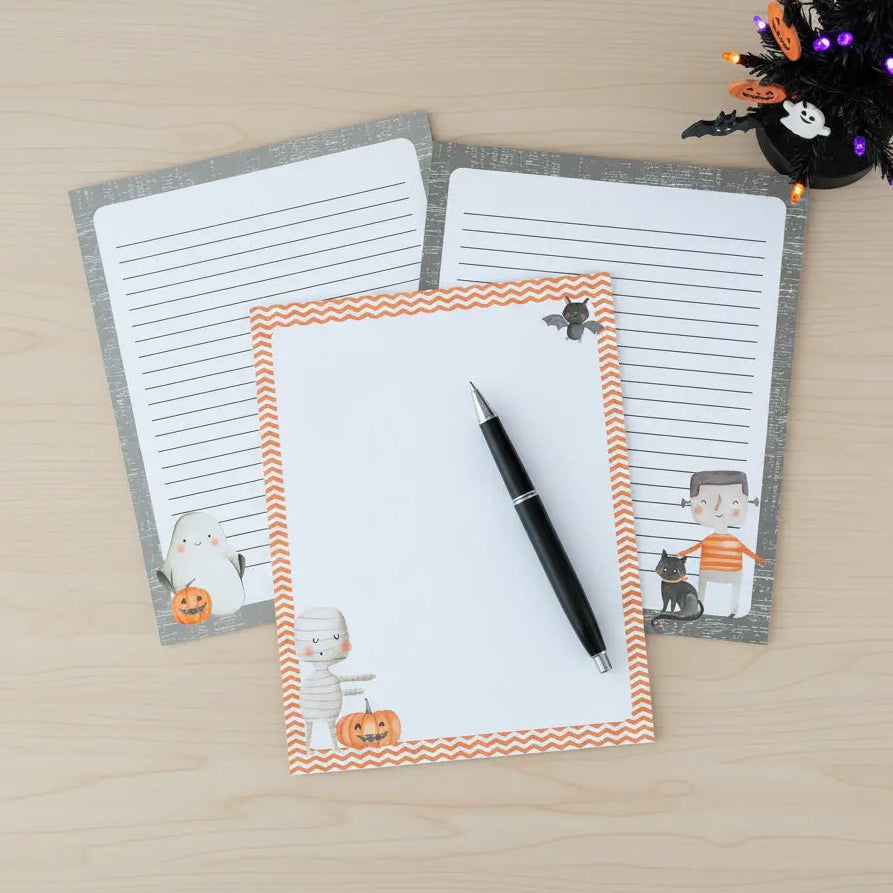 Three Halloween-themed printable papers with a cute ghost, mummy, Frankenstein, pumpkins, and black cat and decorative borders on a wooden surface.