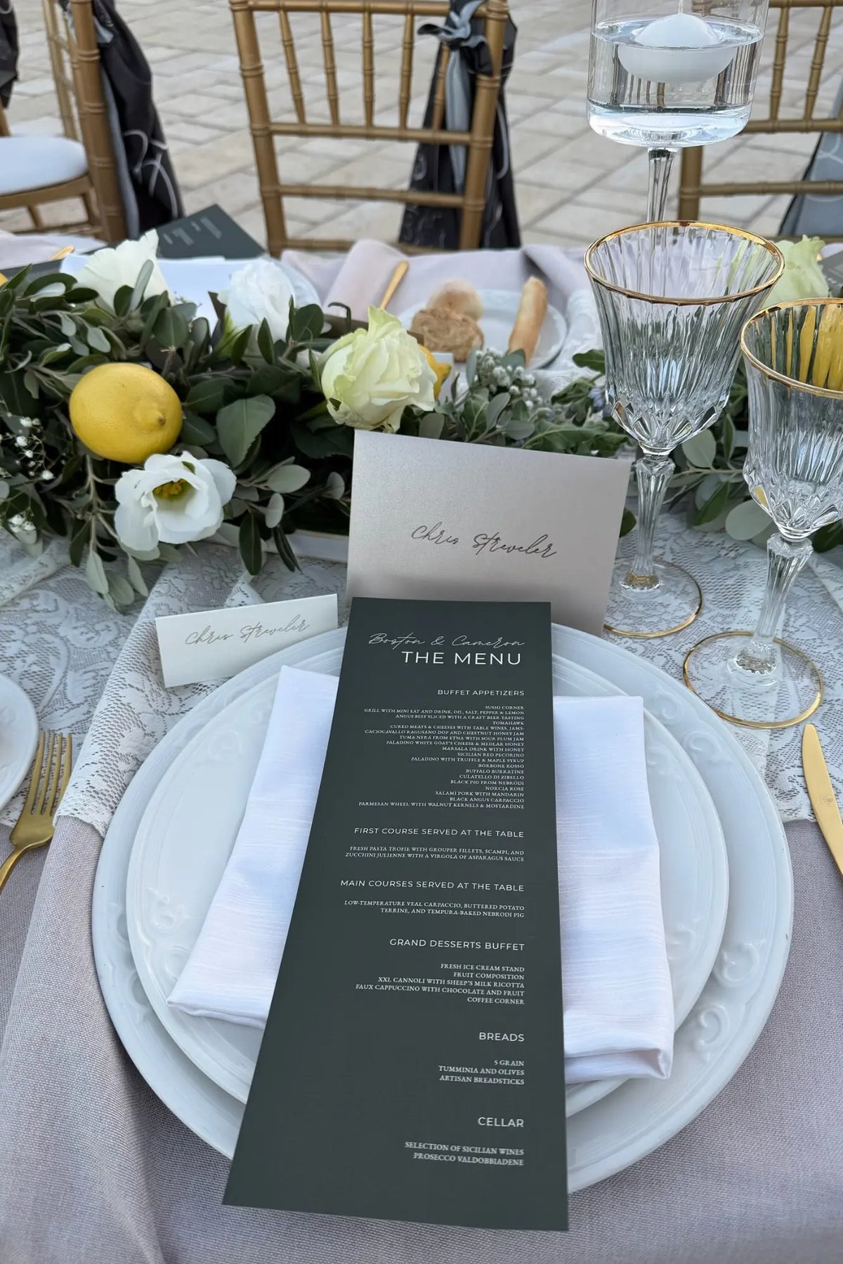 A wedding or event table setting featuring a dark green printed event menu card titled 'The Menu' on a white folded napkin, layered over white plates with a blush pink tablecloth. The setting includes floral centerpieces with white roses and greenery, gold chairs, and clear glass goblets.