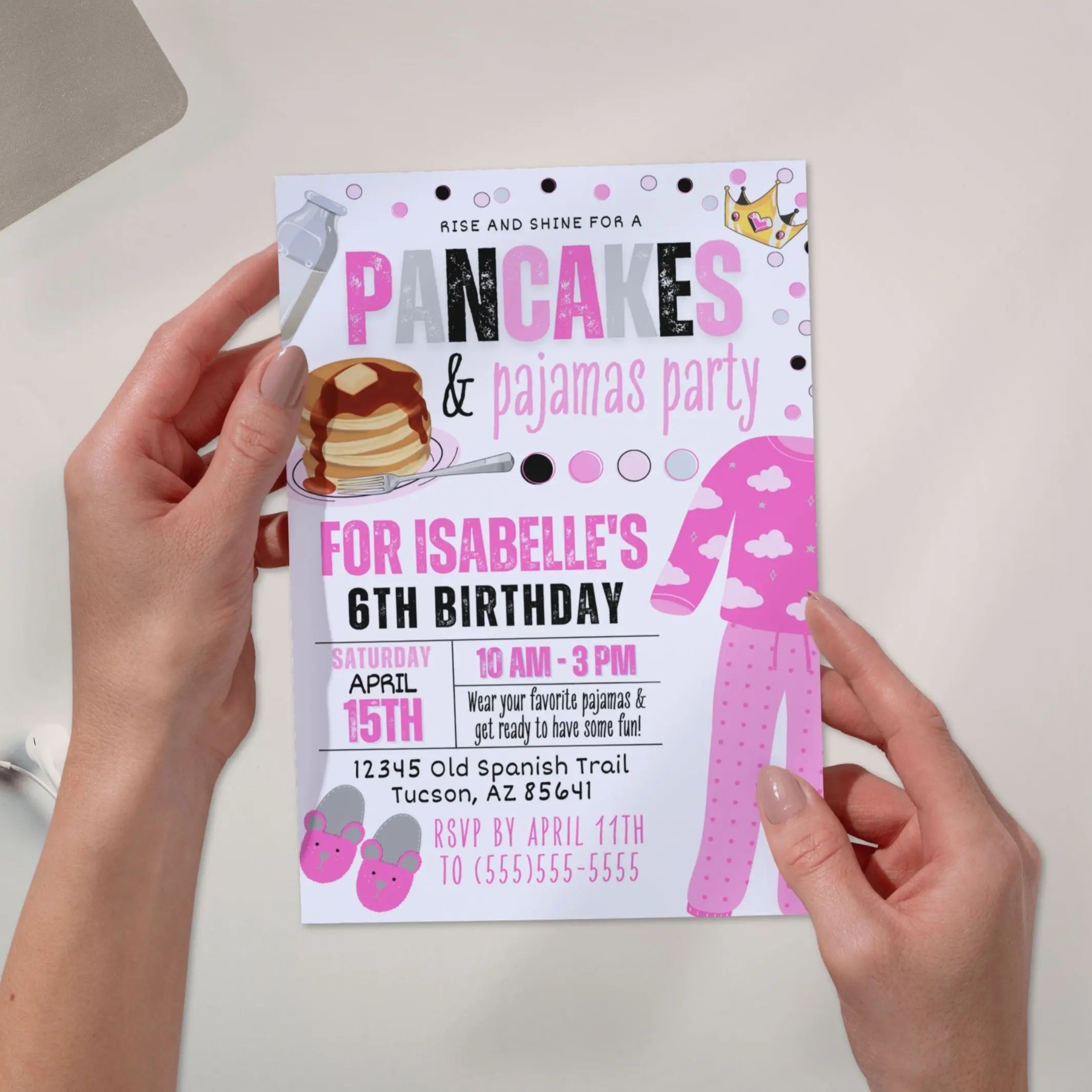 Personalized pink, white and black invitation card for a Pancakes & Pajamas party held by hands on a light background