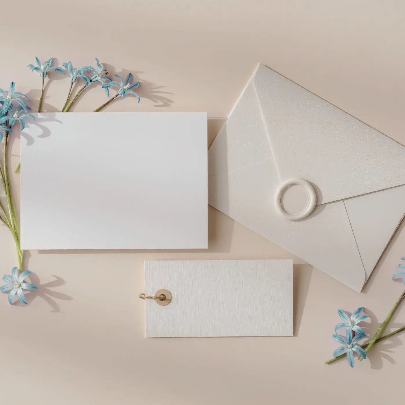 White stationery items including a card, envelope, and tag on a beige background with blue flowers representing a collection of pop by tags and referral thank you cards for small business and real estate agents by Simple Desert Designs.