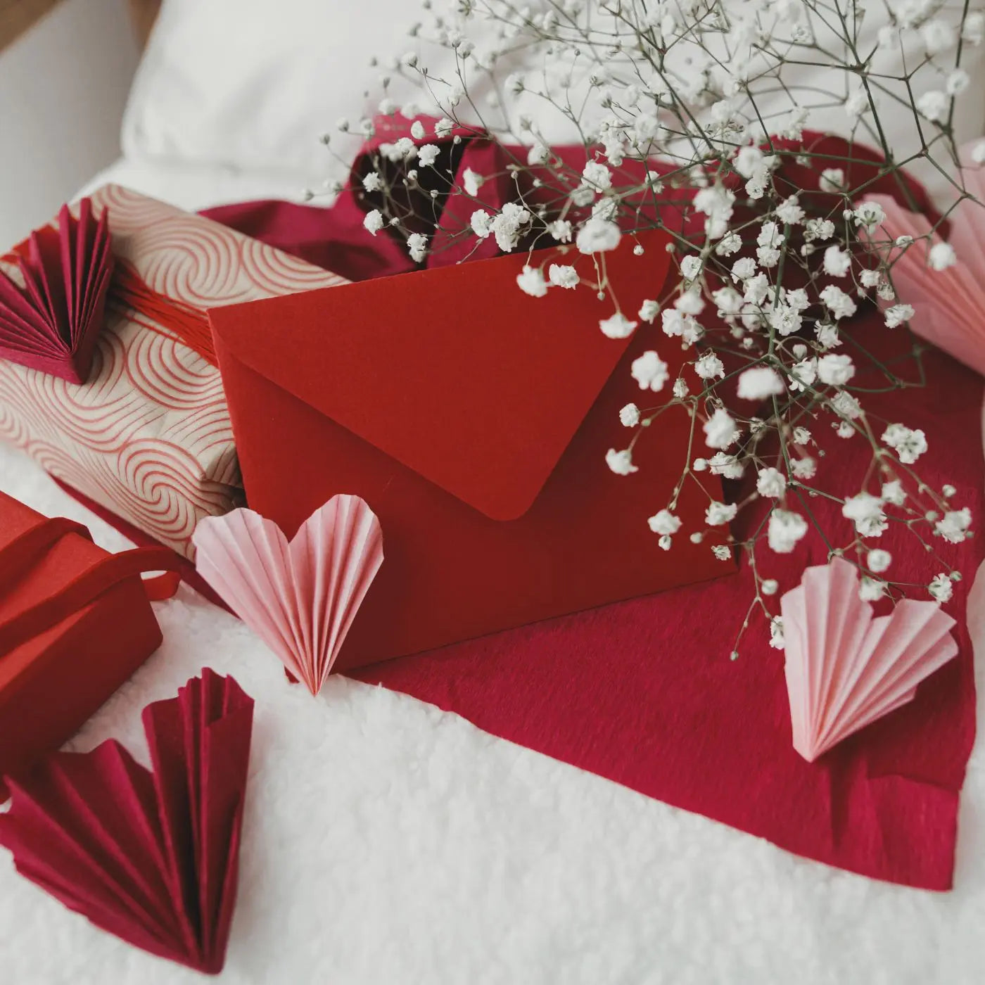 Red envelopes and paper hearts on a white surface with small white flowers representing editable Valentine's Day themed invitations, cards, tags, flyers, and event bundle templates by Simple Desert Designs.